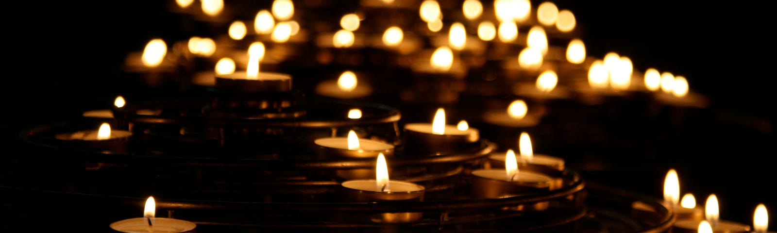 Dim photo of many lit tea lights receding into a black background.