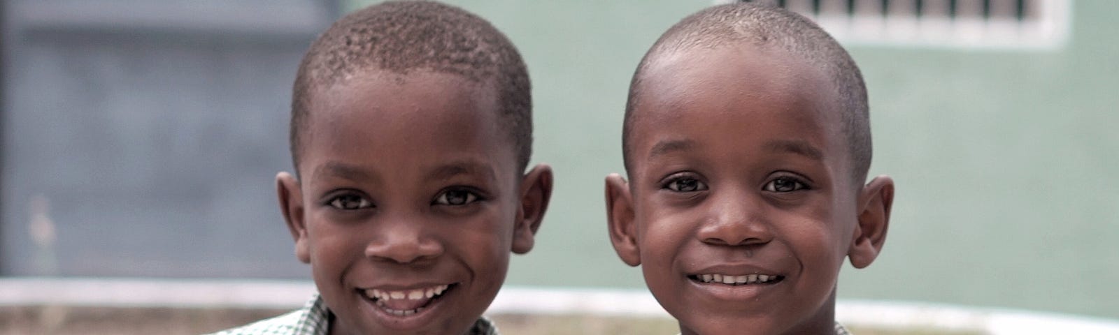The image is of two school aged boys, around 5 or 6; they are in posed in front of what appears to be their school building. They are smiling for the camera in their green checked school shirts.