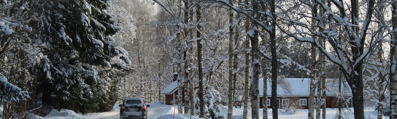 A car drives along a snow-covered country road, lined with trees. A rural building is in the distance.