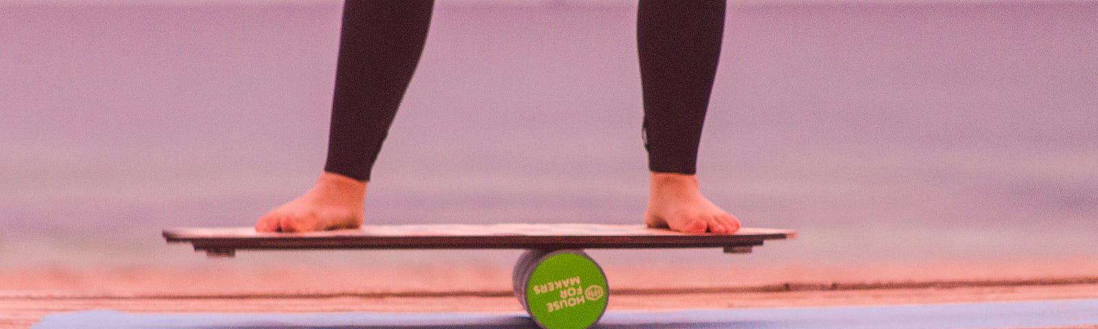 An image of legs from the knees down wearing black leggings and bare feet balancing on a board that is on a round green cylinder.