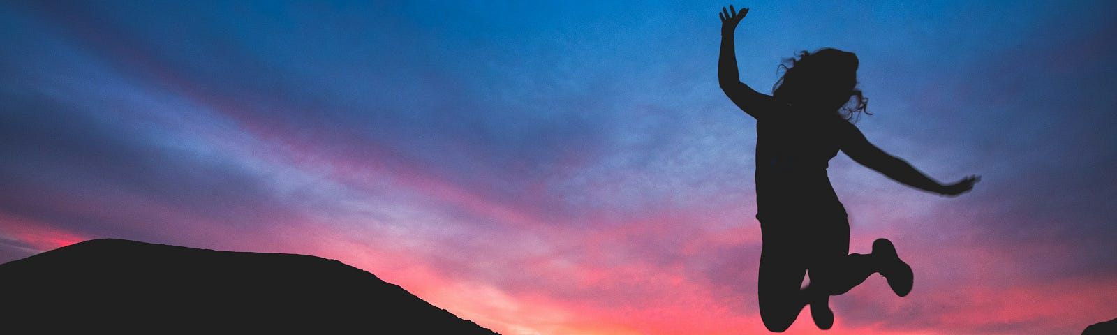 silhouette of female leaping joyfully in the air against a sunset sky and mountains in the background