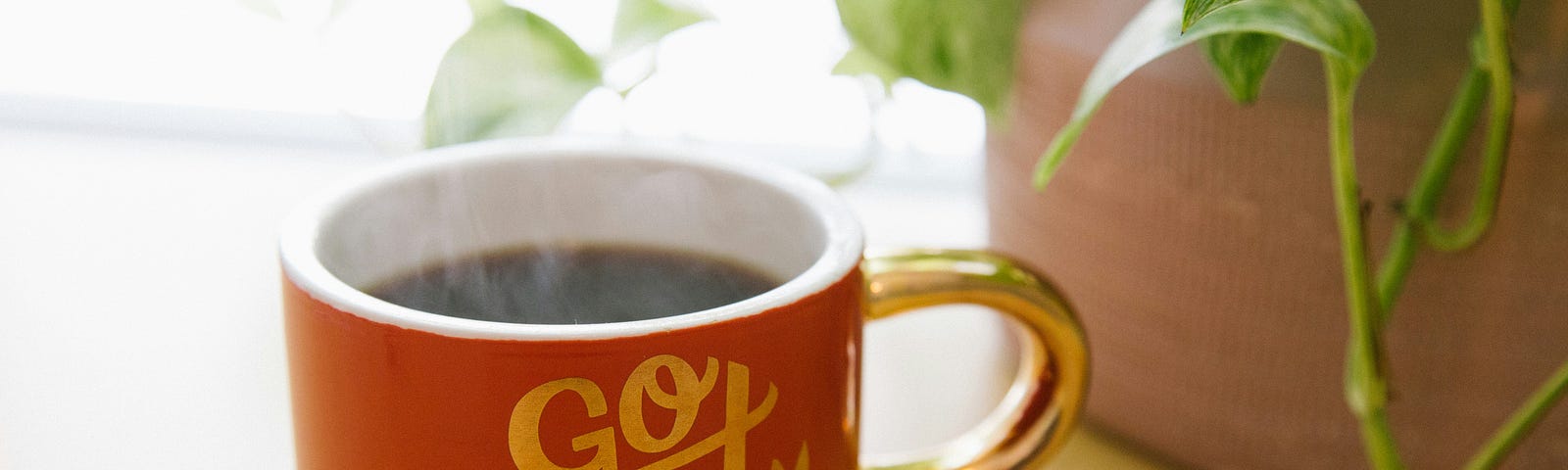 A red mug full of black coffee sitting on a tabletop beside a pot with poison ivy plant