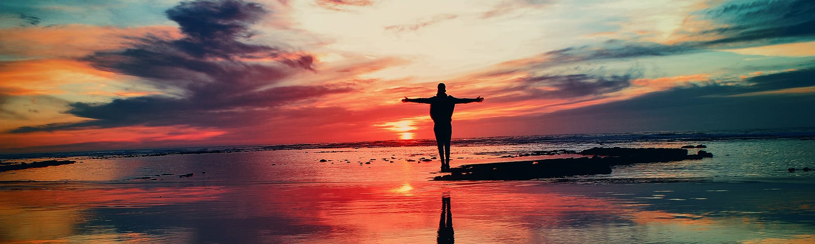 Person standing on a tidal flat with arms spread welcoming the dawn in reflected colours of blues, yellows, pinks, and purples.