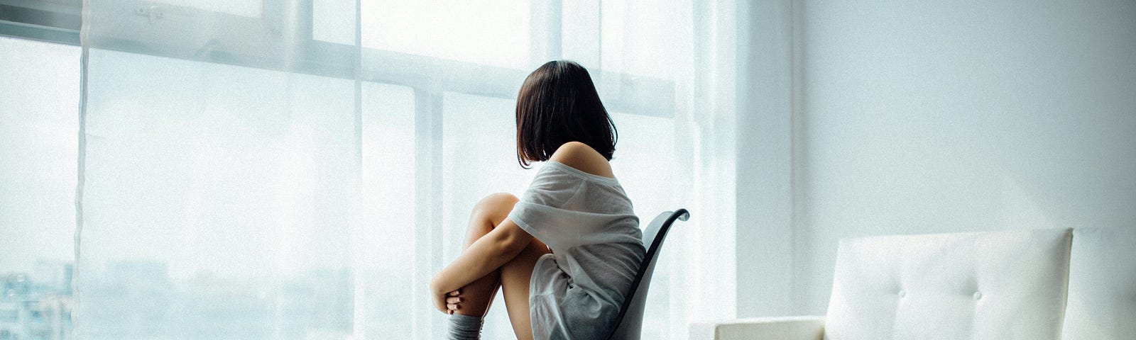 A woman sits on a chair, hugging her knees, staring out the window.