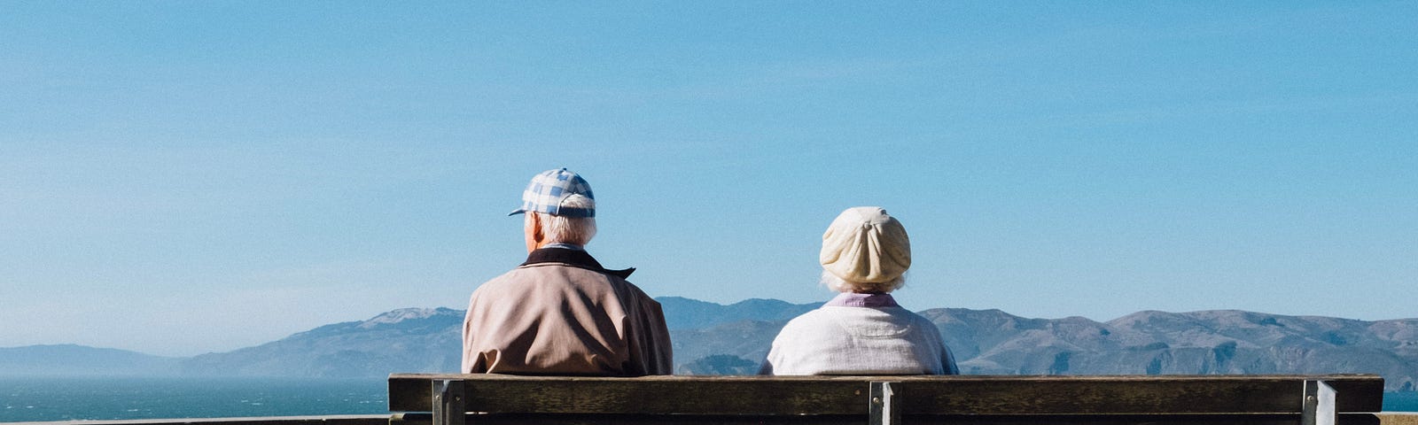 An old couple sits on a wooden bench in silence and looks out at the sky and mountains.