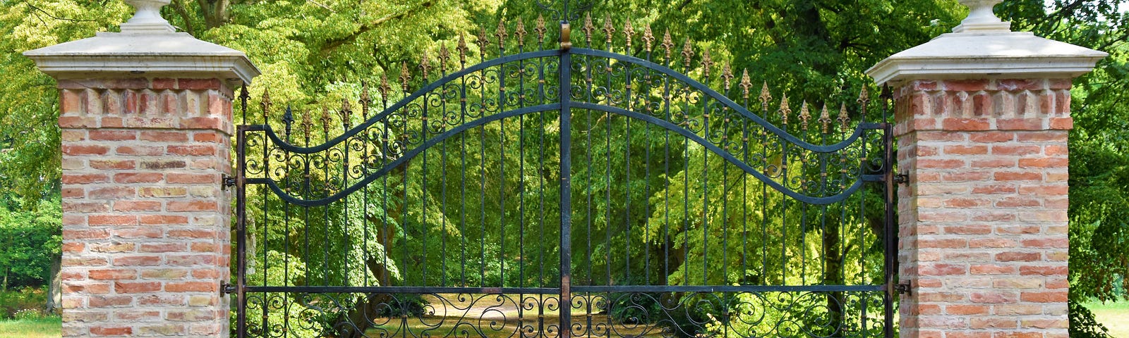 An iron gateway between two columns is closed to a cemetery and green trees behind it.
