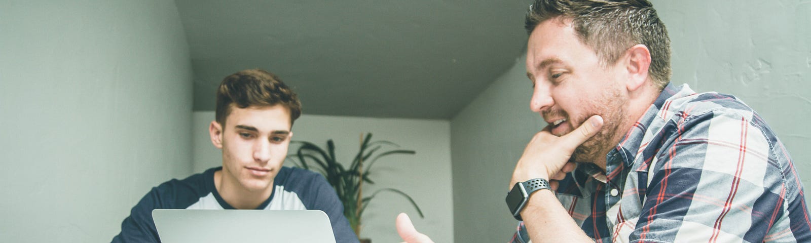 Two men sit and chat at a table while referencing a laptop.