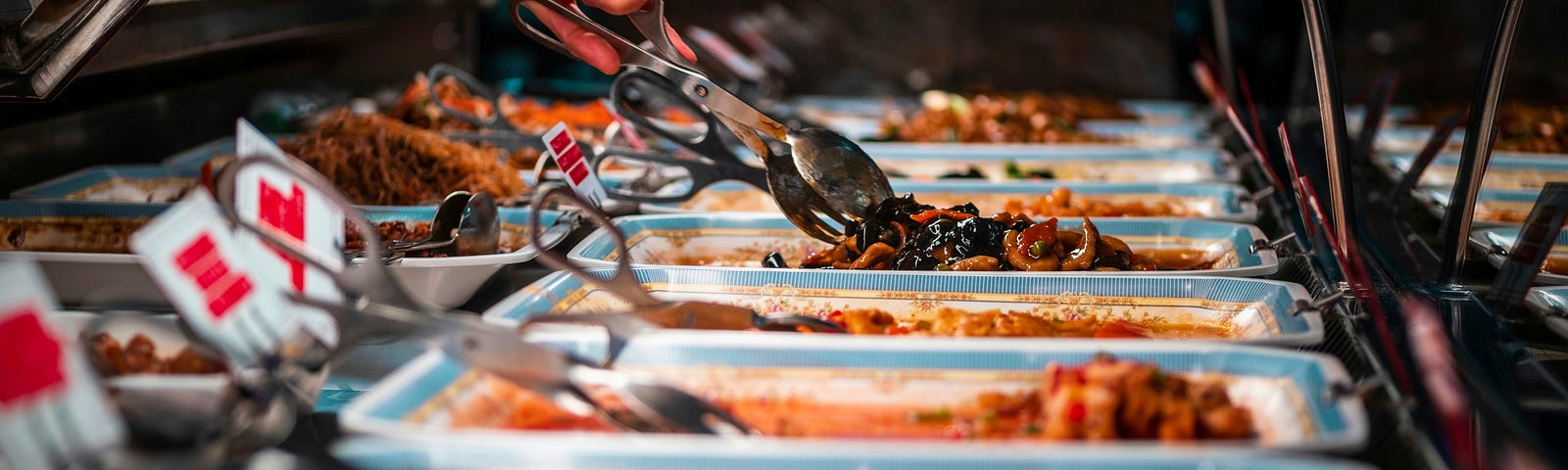 A buffet with various dishes on hotplates. A woman takes some of the food with a spoon.