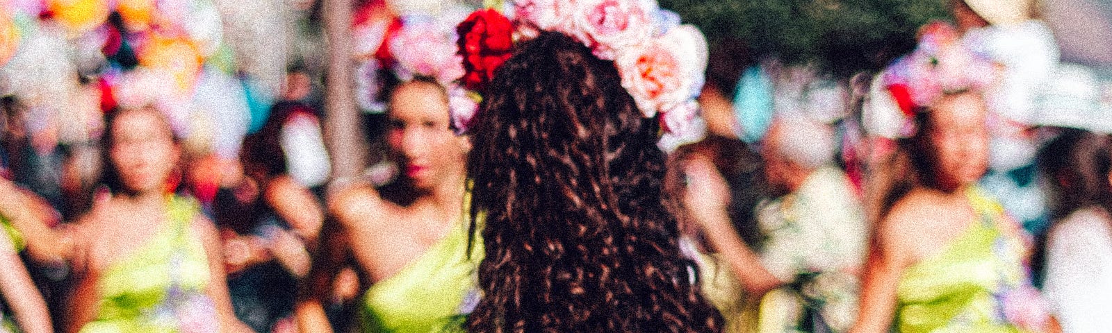 A woman in a lime green dress is in the foreground while several other women dressed the same are in the background. The woman in the foreground has curly black hair with a flower headdress.