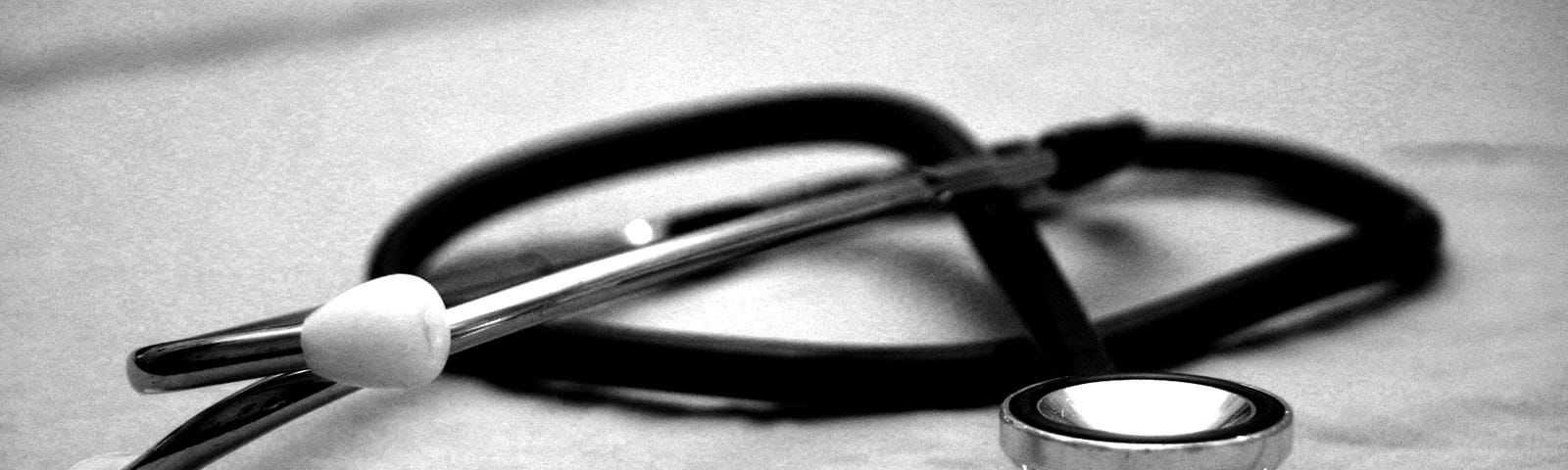 The image shows a black-and-white photograph of a stethoscope lying on a cloth surface, possibly a hospital bed or examination table. The stethoscope is positioned with the chest piece facing upward and the ear tips resting on the cloth. The focus of the image is on the chest piece, which is in the foreground, while the ear tips and tubing extend into the background. The overall mood of the image is clinical and professional, evoking the atmosphere of a medical environment.