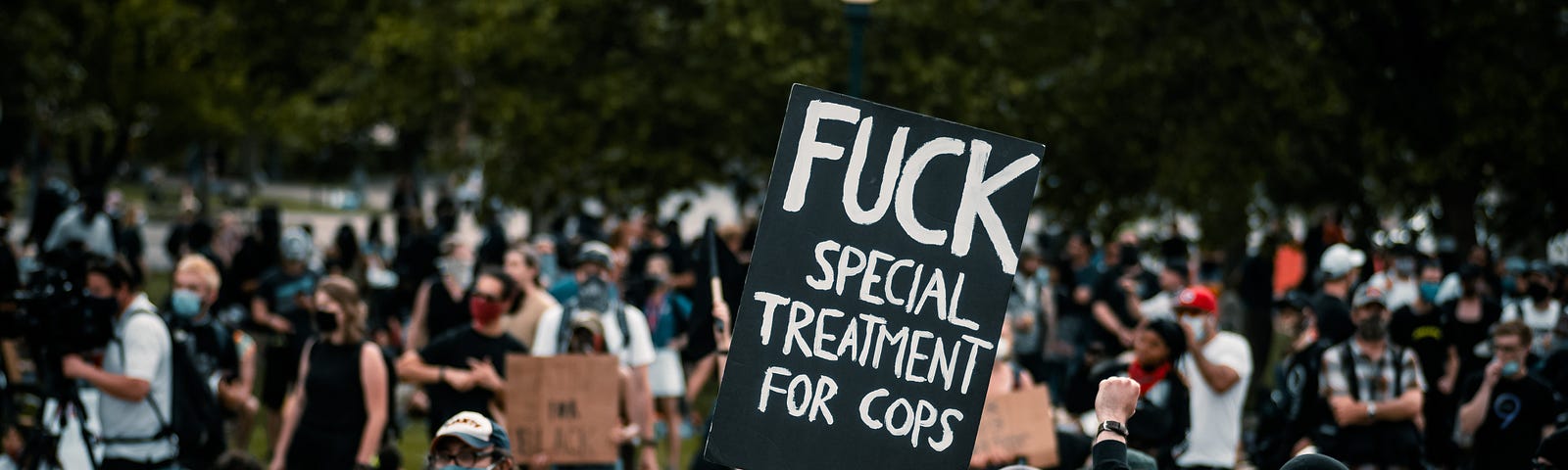 A man standing in a crowd protesting at a park holds up a black sign with white text that reads: Fuck special treatment for cops