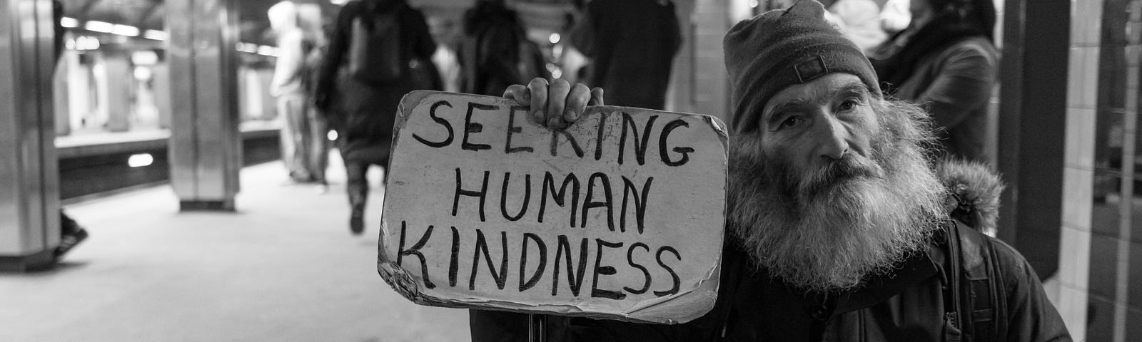 An old man with a beard on a subway station platform holding a sign that reads “Seeking Human Kindness.”