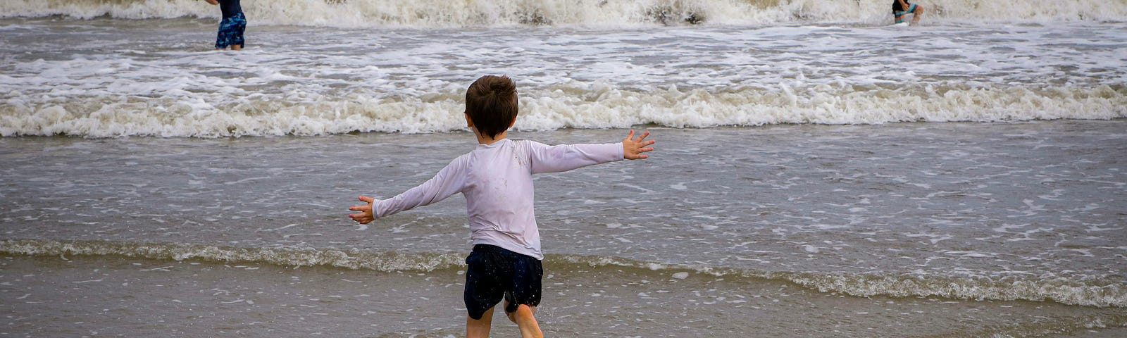 young boy in white top and black shorts running towards the sea with his arms outstretched while two more children can be seen in the waves