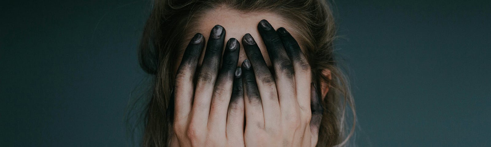 A young woman covering her face with both hands — her fingertips are black, they look singed and burnt.