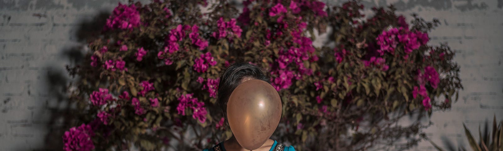 A woman standing in front of a wall of flowers with her face hidden by a balloon.