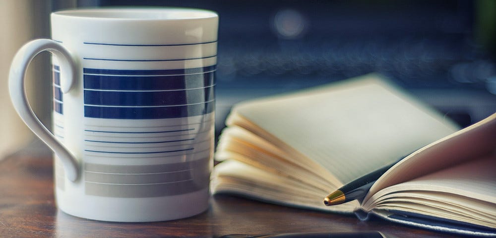 A coffee mug sits beside an open notebook with a pen resting in it, all on a wooden desk.