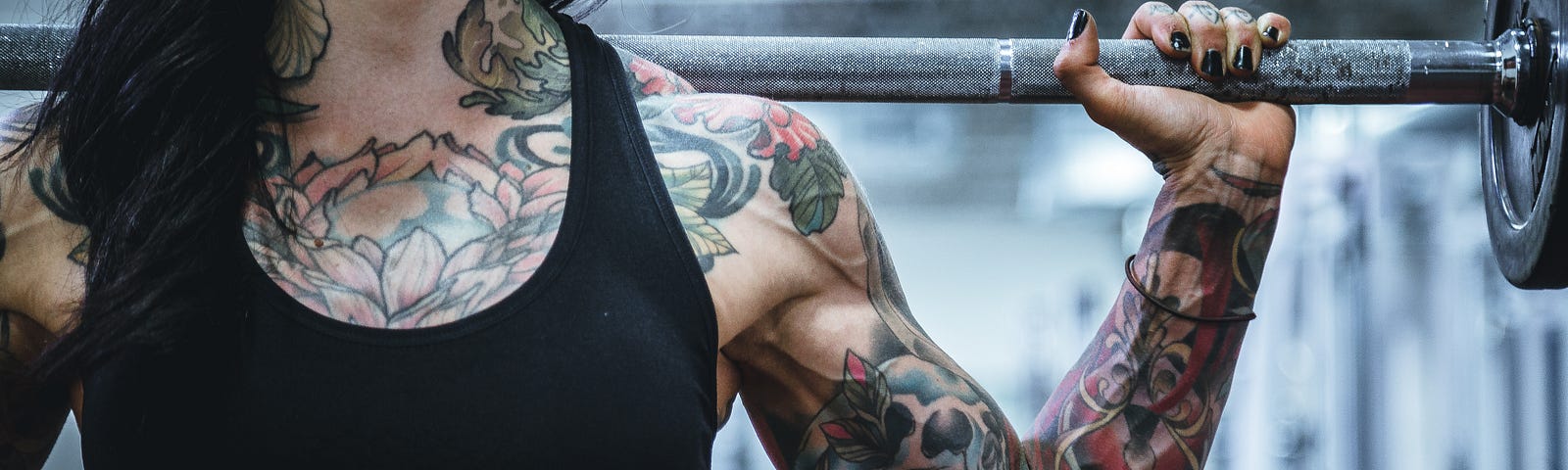 A heavily tattooed and brunette women faces us, waist to chin level, as she pushes up a barbell behind her in a shoulder press. Blurred background.