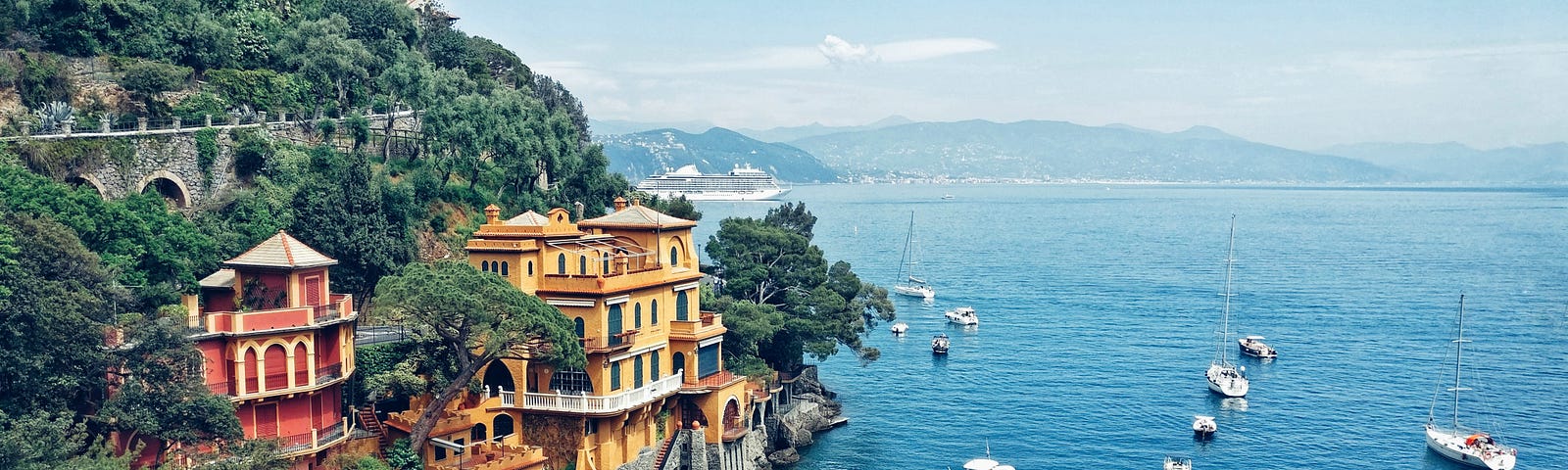 Large yellow mansion perched on a cliff overlooking an inlet on the sea near Portofino. There are many small boats moored and mountains can be seen in the distance.