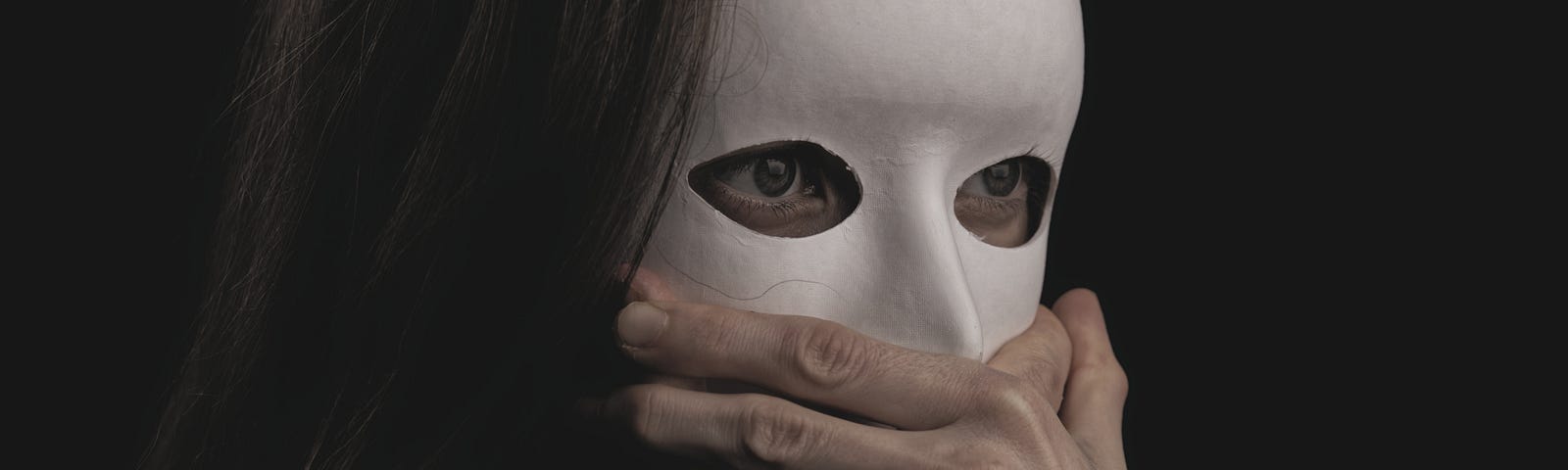 A young brunette woman with long hair is wearing a white cardboard mask, hiding her entire face. But her eyes show through cut out eye holes. They are staring widely at something off-camera. Two unidentifiable hands are grasping where her mouth ought to be, stopping her from speaking. Allodoxaphobia: the fear of hearing others’ opinions. My phobia stopped me from speaking out against child abuse. I was unable to confront an abusive parent in public. What would you have done in my situation?