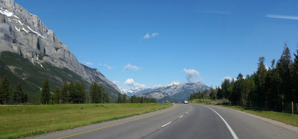 Road curving between green verges and trees, towards the rocky mountains in the background, under a blue sky with a few clouds.