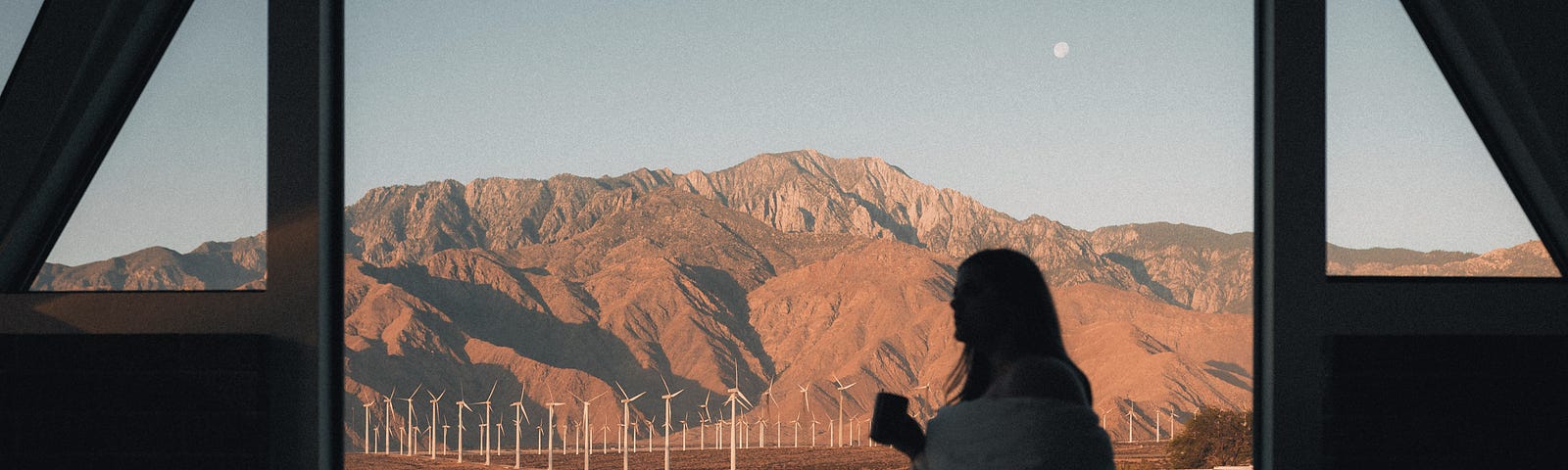 A woman sits in the shadows inside a cabin with floor to ceiling windows. She looks outside with a blanket wrapped around her and a mug in her hand. Through the windows are fields of wind mills against the Palm Springs desert mountains at sunrise. The moon is visible and the sky is gray.