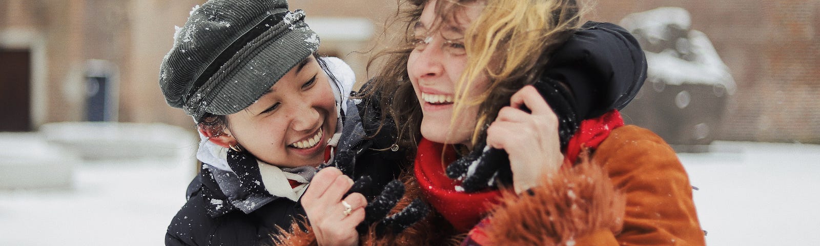 A young woman in a dark jacket and cap stands close to another woman in an orange jacket. They are laughing as if they are playing together in the snow.