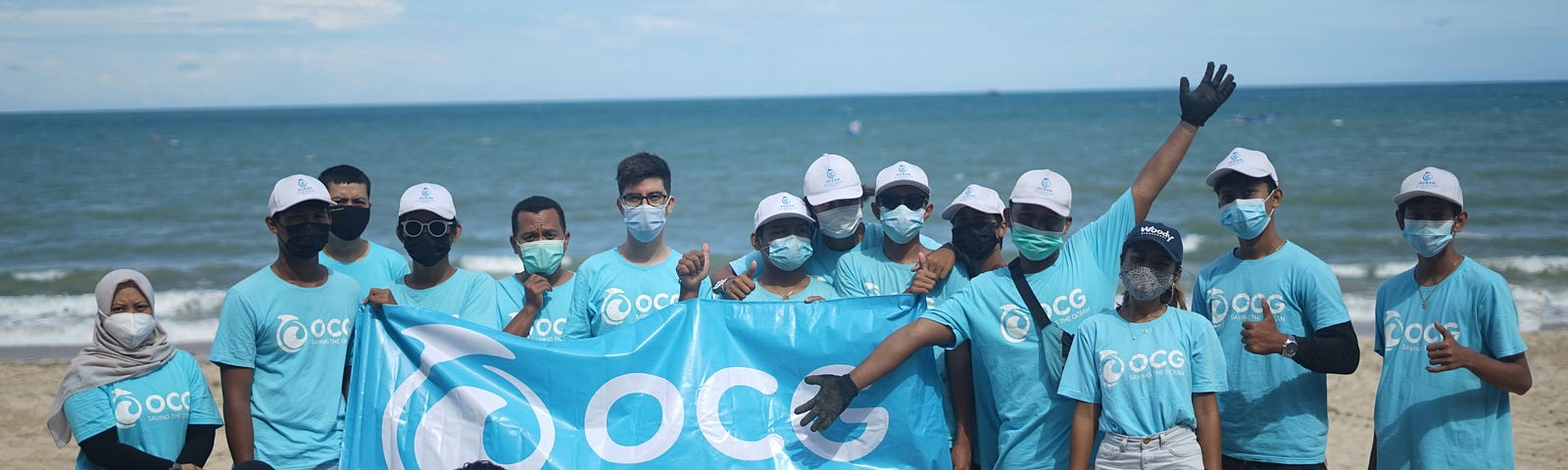 mixed nationality group of people wearing sky blue shirts, collecting beach trash, and holding a banner that says Saving the Ocean