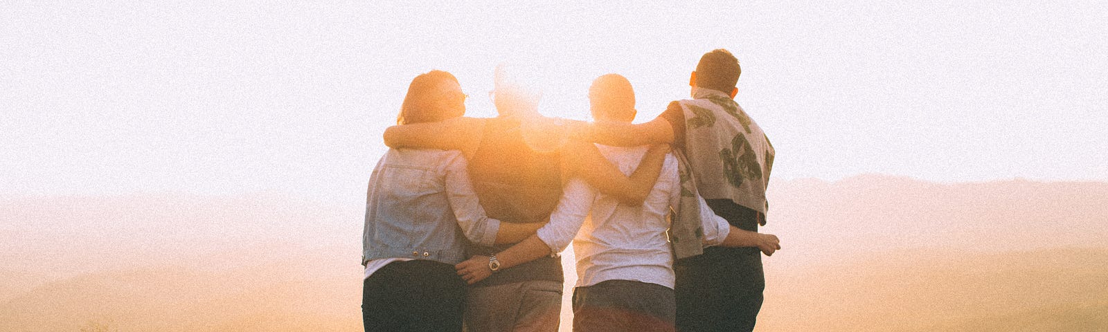 photo of four people standing with their arms around each other’s shoulders, looking away from the camera at rolling hills.