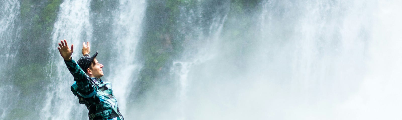Male standing at the side of a waterfall wearing a cap, sporty clothes, arms up in the air, he looks happy.