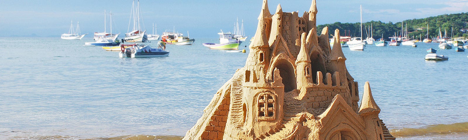 An ornate sandcastle be the sea. Boats are on the bay.
