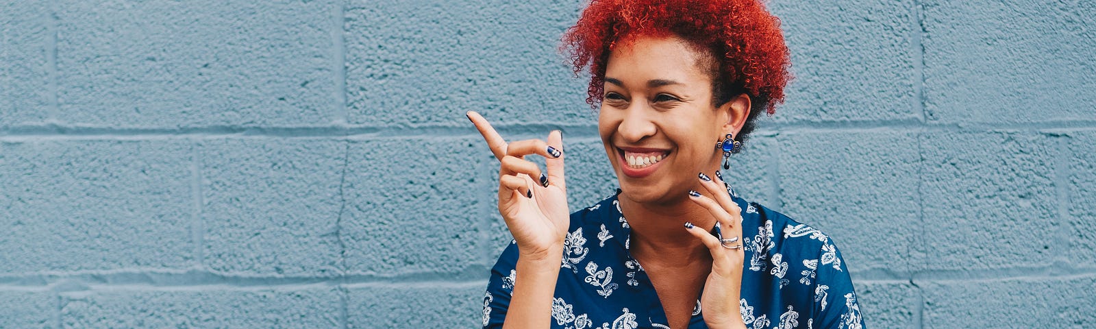 A black woman smiles and looks to her right. She points a finger, wears a blue blouse, and has short curly read hair.