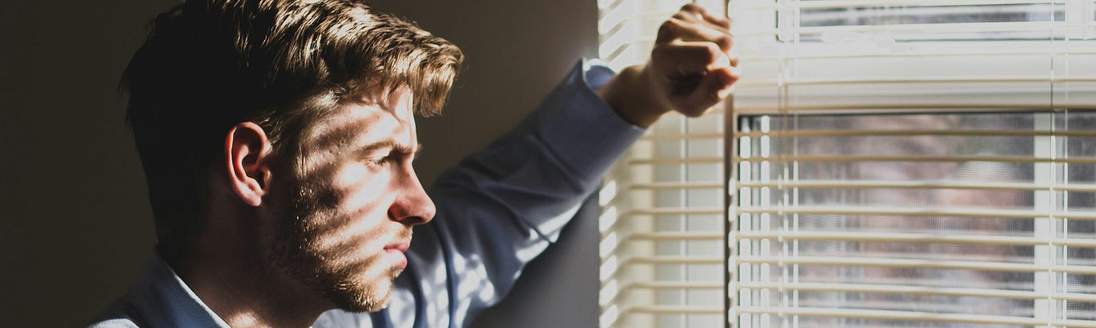 A contemplative man gazes out of a sunlit window, blinds casting striped shadows across his face and shirt.