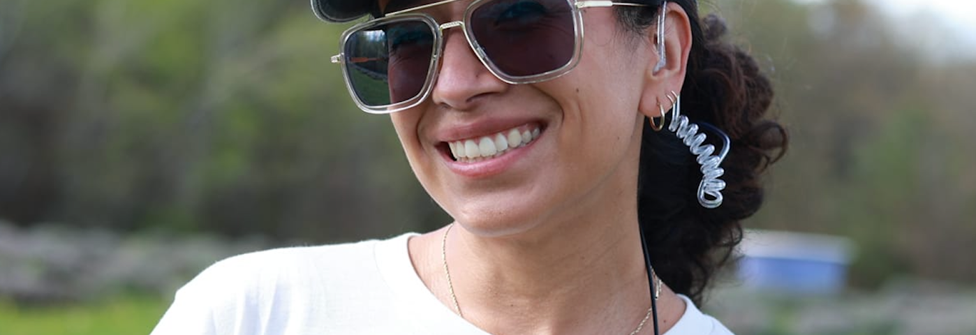 Photo of Emily Baeza smiling at the camera. She is in a green pasture, wearing a white shirt with horror characters which reads “Horror is Queer.”