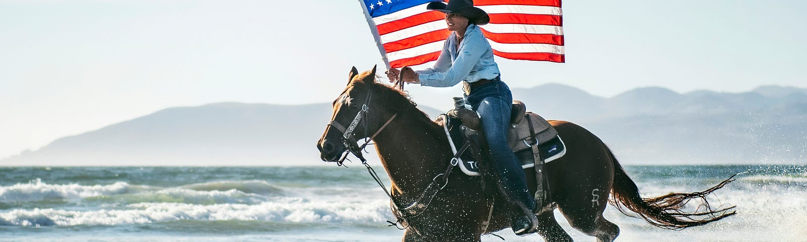 Photo of a person on a horse galloping on the beach with an American flag.