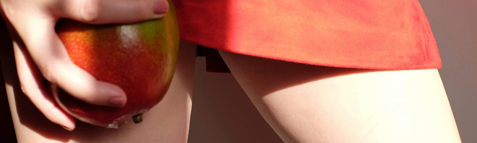 Someone in a reddish skirt is holding a reddish-greenish mango next to a leg.