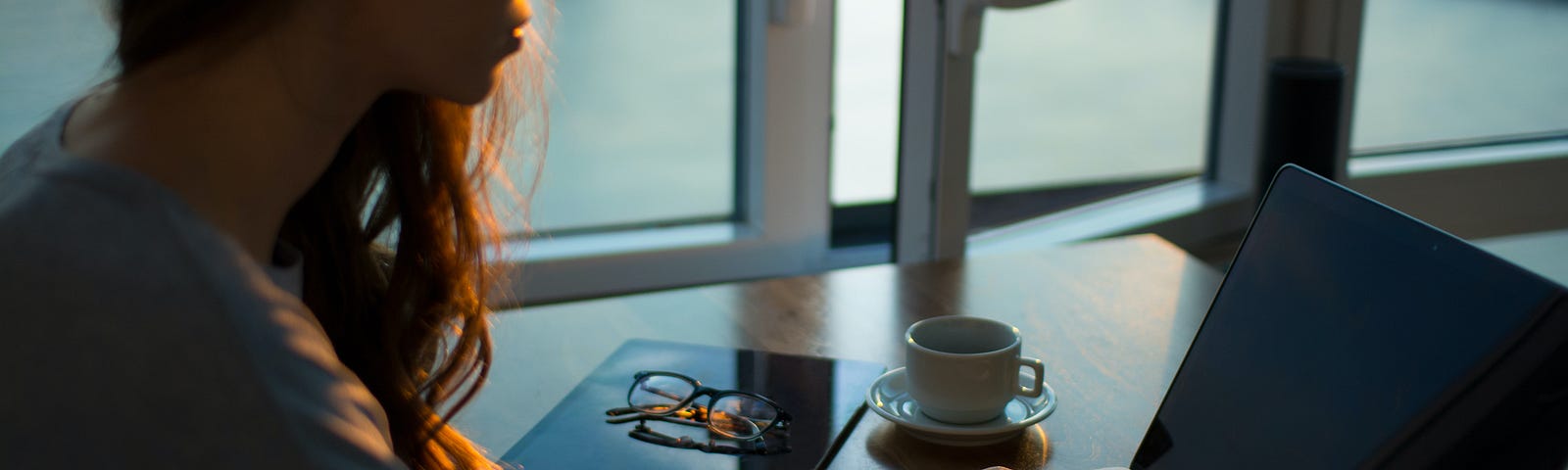 A thoughtful lady with long reddish-brown hair typing on a laptop in front of a partially open window. Other writing accessories and a coffee cup surround the laptop.