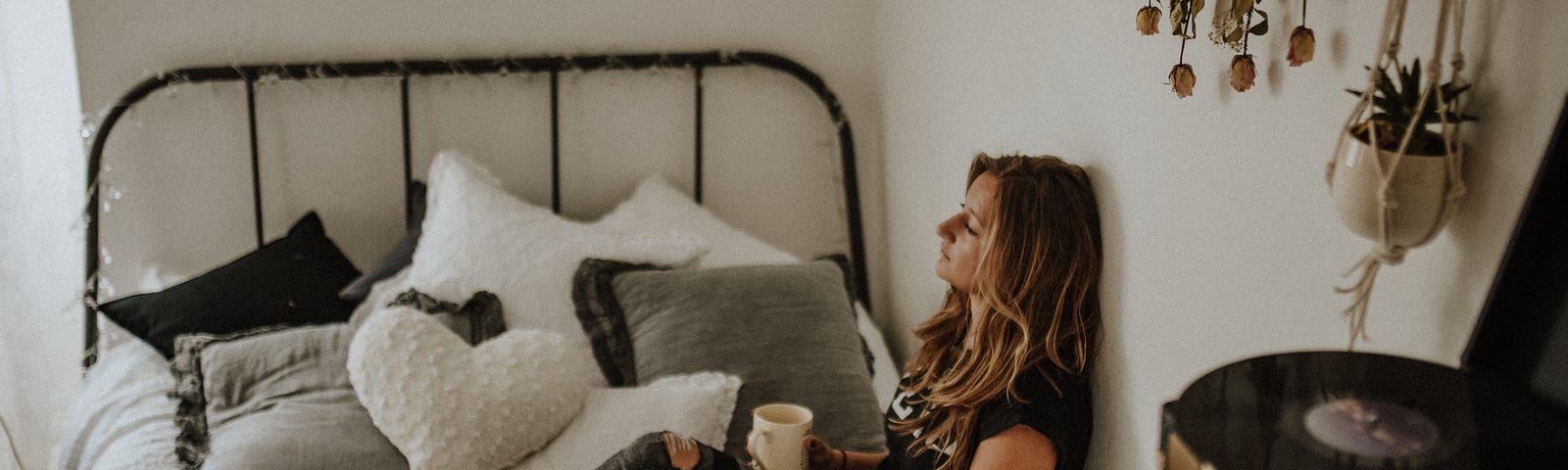 A white woman, wearing a black shirt and jeans, sits on her bed, holding a notebook and cup of coffee. She is gazing out the window. On the bed are grey and white cushions. On the wall behind her is some plant decorations. Next to the bed is a crate with a vintage record player on it.