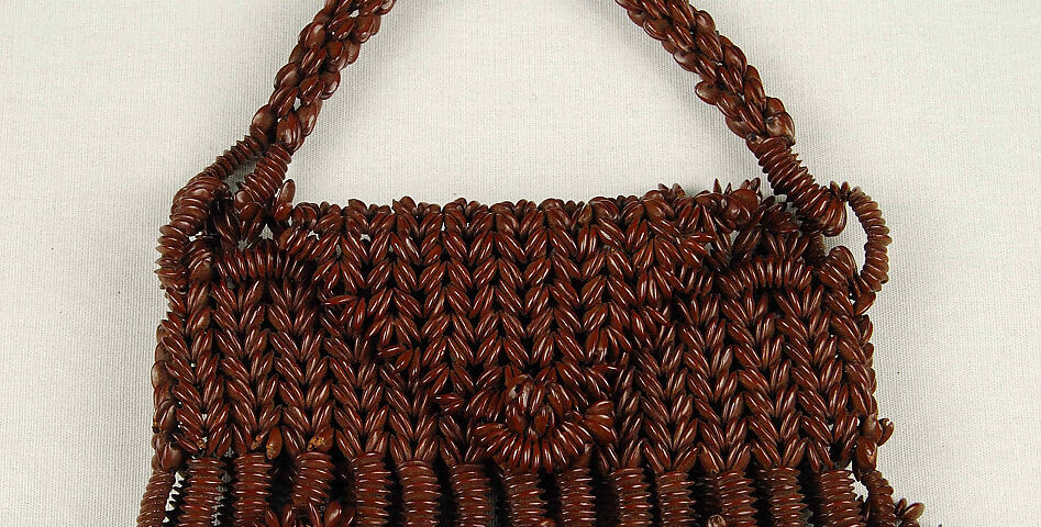 A photograph of a handbag with a short strap and a triangle shaped flap to close the top. The outside of the handbag is made using tamarind seeds and designs with tamarind seeds. The handbag is placed on a light background.