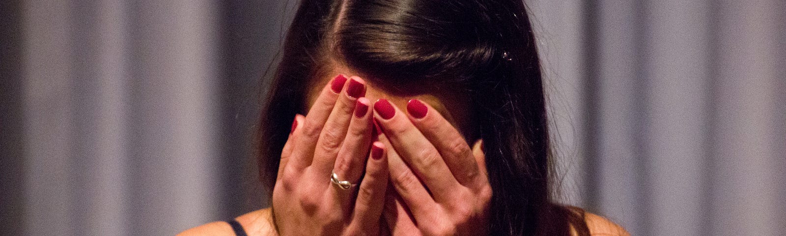 woman with long dark hair, hands with red nails hiding her face