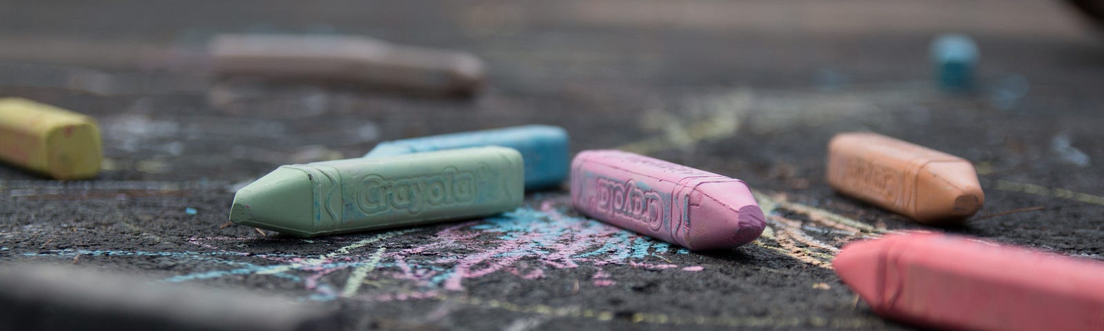 A close-up asphalt ground view of a bunch of crayon-shaped chalks of different colors left strewn on the ground over some of their scribbles.