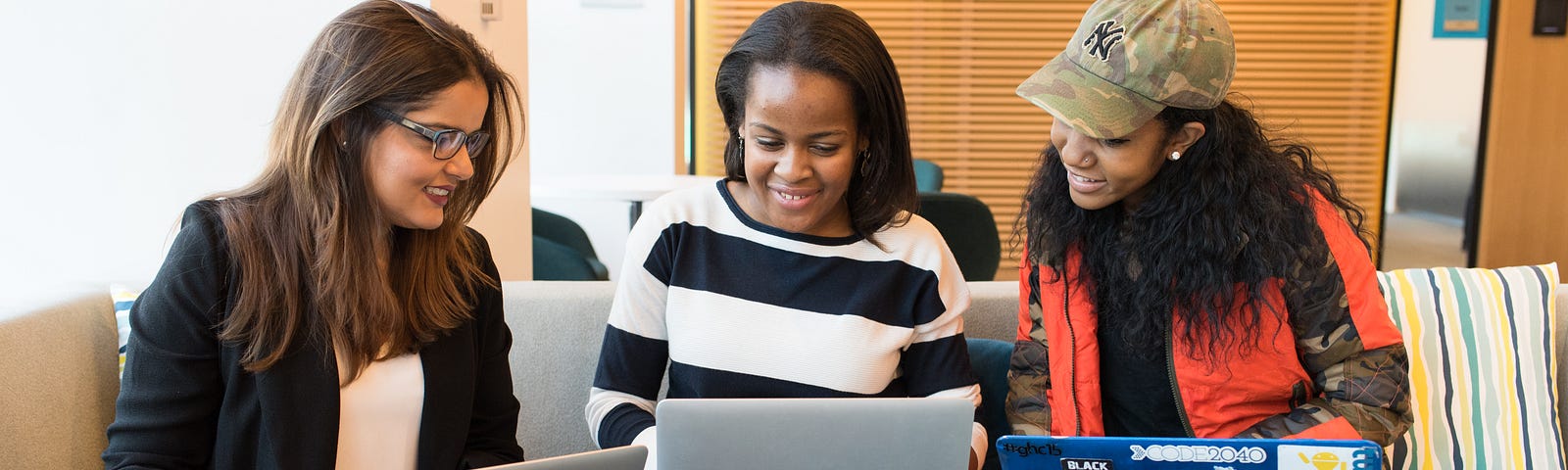 Three women sitting next to other, smiling, and looking at the middle woman’s laptop (all three have a laptop)