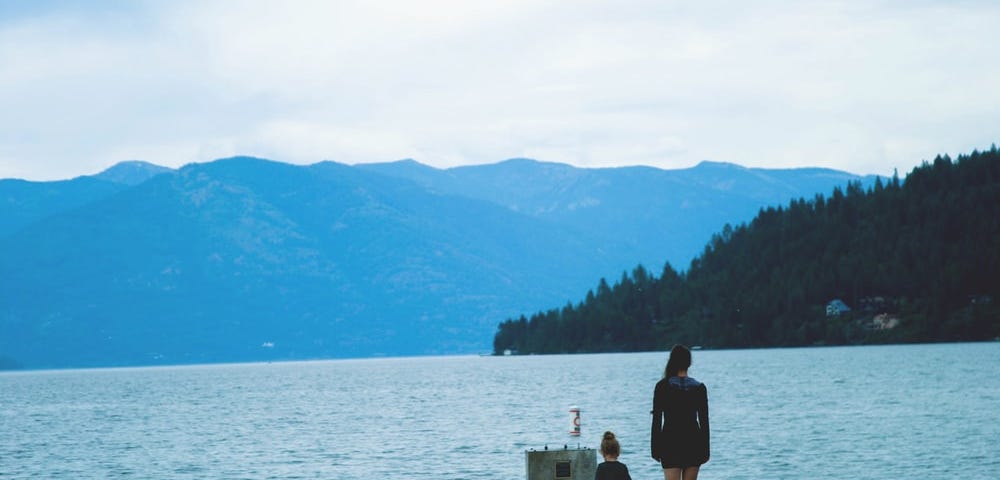 A woman and little girl walking side by side on a pier on the sea. Mountains and trees are visible in the near distance.