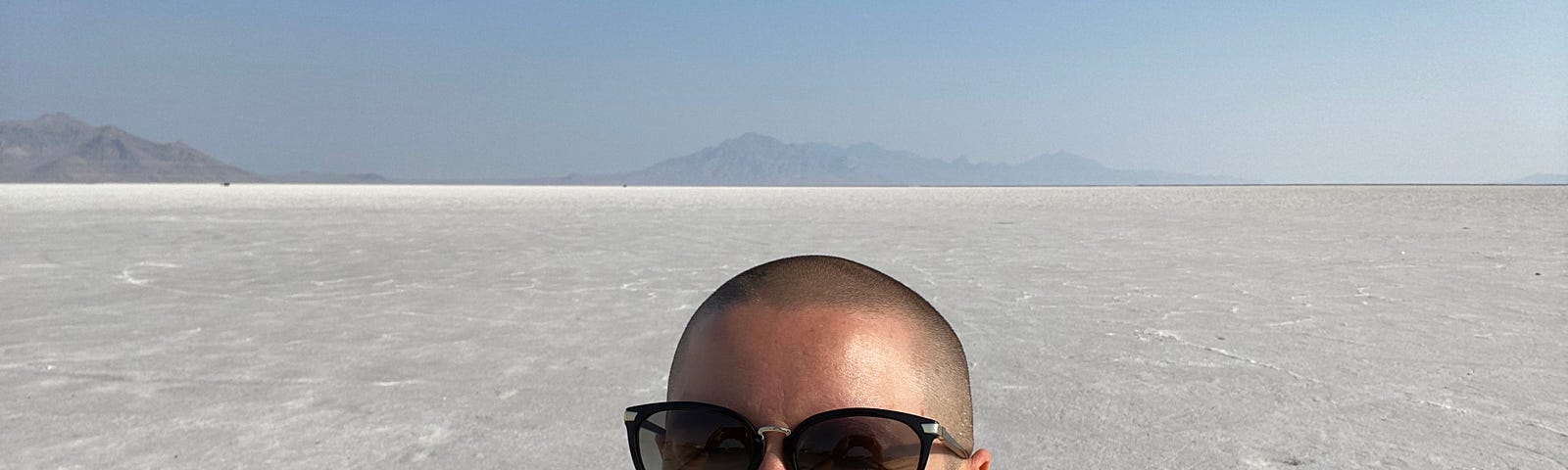 Sarah Dopp smiling with a large stretch of salt flats in the background. She is white, has shaved head, wearing sunglasses.