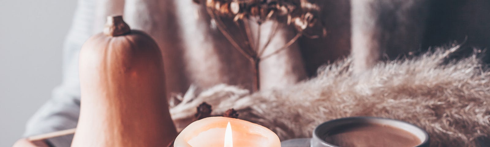 A cowled figure holds a small ceramic tray. It contains a mug of hot chocolate, a lit candle, a butternut squash and a spray of dried branches and flowers.