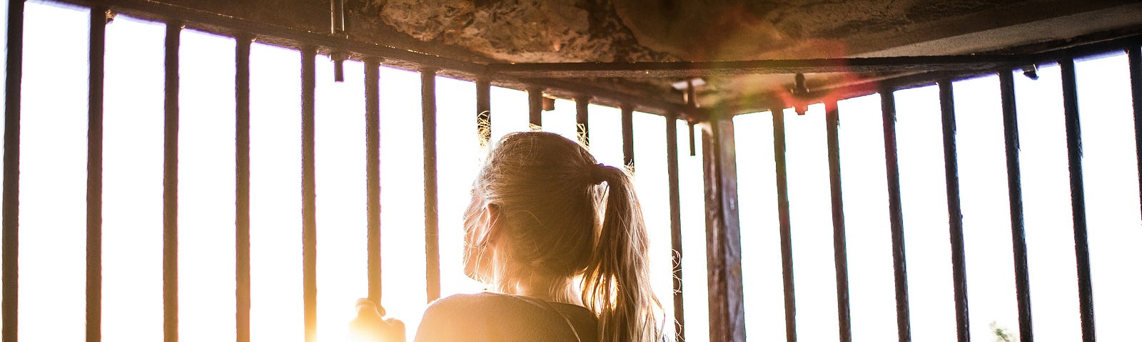 A woman stands behind bars looking out at the world. Self-imprisoned, she is insecure about life beyond the confines of walls. Light peers in through a grey sky warming her, making her think of all she is missing. She wear a dark sweatshirt and sports a long blonde pony tail.