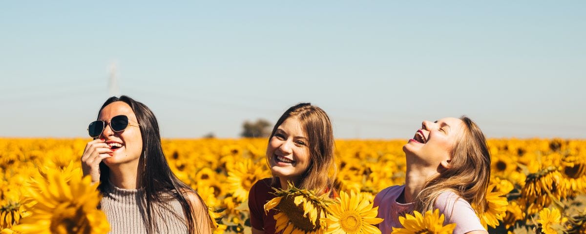 Three women with long hair laughing in a field of sunflowers to boost assertive communication style by Suzanne Marie