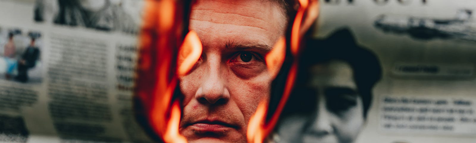 A man’s face is seem through a burning sheet of newspaper.