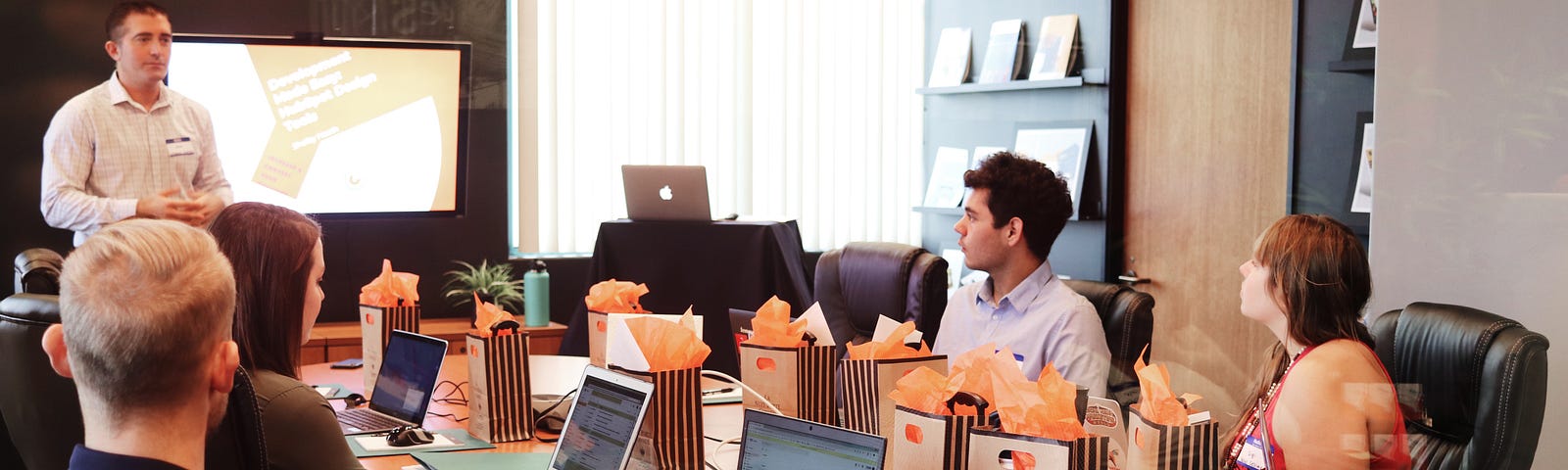 Team of 5 people sitting in an office board room with laptops, doing training with a presenter.