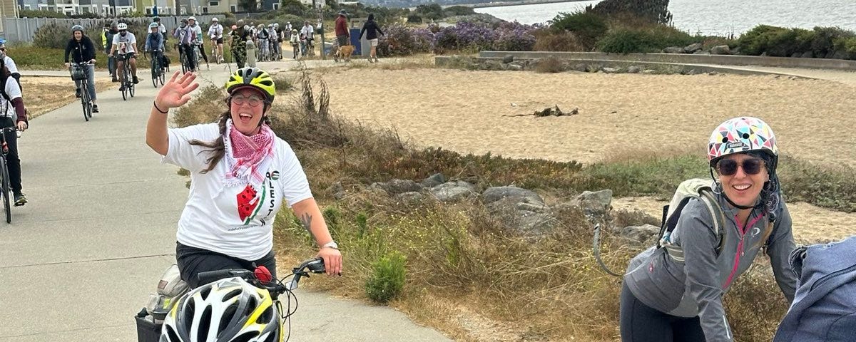 A view along the Bay Trail with three bicyclists from SURJ BA in front and many others following.