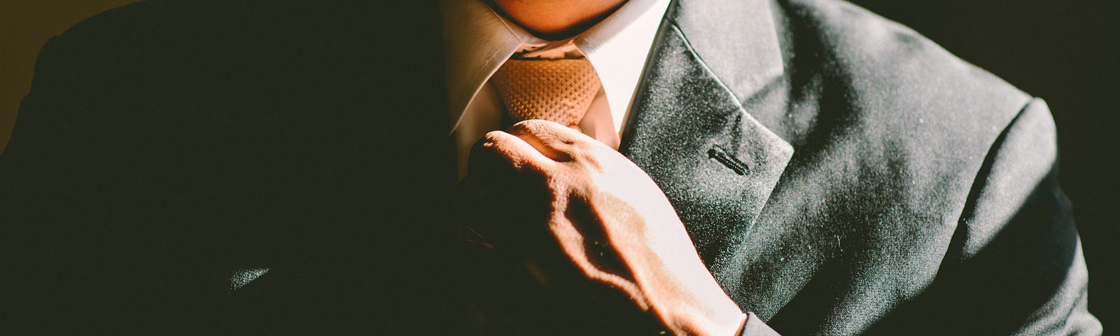 Photo of a man wearing a grey suit and adjusting his tie. The photo has been cropped to show just his chest and neck.